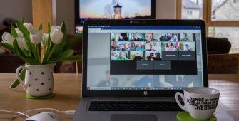 Ein Bild von einem Laptop, auf dem mehrere Personen in einem Online-Meeting sind. Es isnd eineige Personen mit Bild und in einigen Feldern steht ein Name. Rechts neben dem Laptop steht eine weiße Kaffeetasse, links daneben eine Vase mit weißen Tulpen. Links vor dem Laptop ist eine weißen Computermaus.